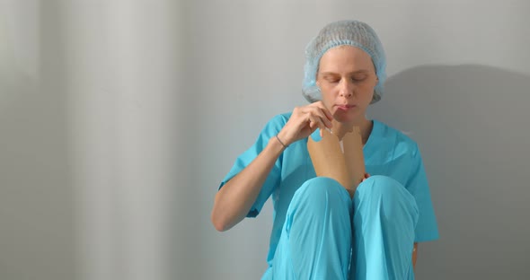Woman Nurse Eating Takeaway Food From Disposable Box Sitting on Floor at Hospital alt