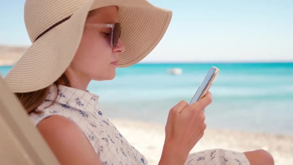 Woman in Hat Uses Smartphone App Relaxing on Exotic Sea Beach in Summer Day