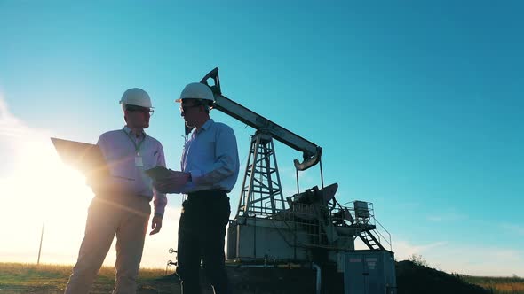 Oil Field Workers Having a Discussion By an Oil Pumpjack in Sunlight alt