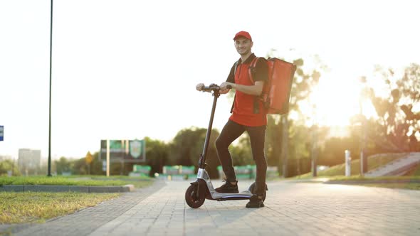 Portrait Deliverman Standing With Electric Scooter With Red Thermal Backpack Looking at Camera