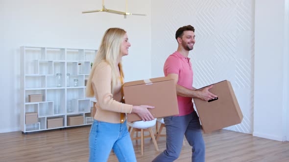 Amazed couple man and lady walk along new apartment room holding large cardboard boxes alt