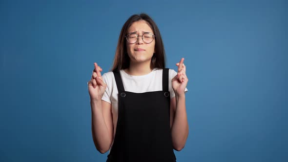 Cute Asian Young Girl Praying Over Blue Background. Woman in Glasses Begging Someone. . alt