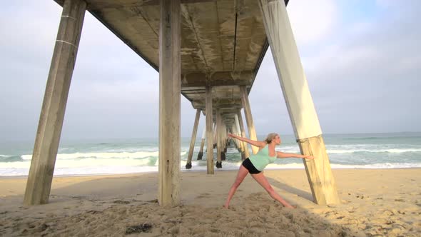 A young attractive woman doing yoga on the beach under a pier. alt