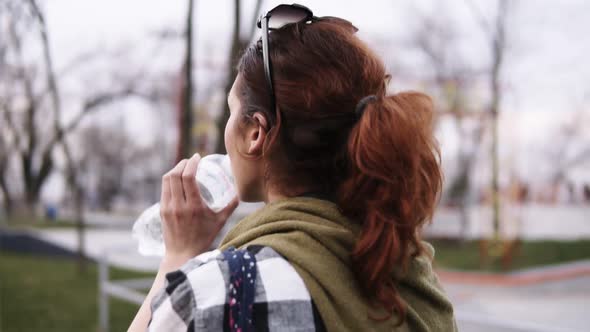 A Girl with a Tail Walks on an Overcast Day in the Park Drinks Water From a Plastic Bottle alt