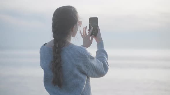 Stylish Young Woman in Sunglasses Shoot Sea on Phone at Sunset Alone on Coast