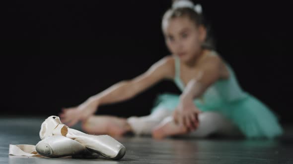 Teenage Ballerina Girl in Blue Tutu Sits on Floor Stretching Ballet Shoes Pointe Shoes in Foreground alt