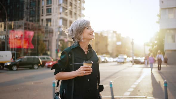 Middle Age Woman Goes Through the City with Coffee in Her Hands and Smiles alt