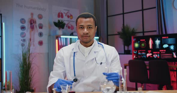 African American Chemist in white Coat posing on camera with glass flask and test tubes filling alt