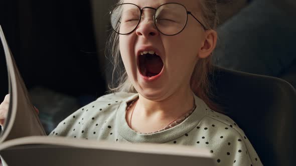 Little child tired girl reading a book in glasses after school and yawns alt