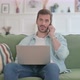 Young Man with Laptop Talking on Phone on Sofa