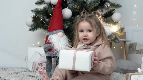 Sweet Little Girl Sitting Near Christmas Tree with a Present Box in Her Hands alt