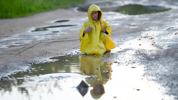 Little Cute Girl in a Yellow Raincoat Plays and Throws Stones in the Puddle on the Road