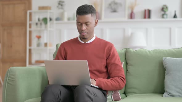 African Man Smiling at Camera while using Laptop on Sofa alt