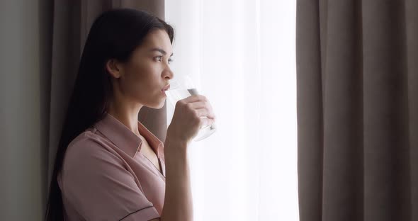Woman Stands Near the Window with the Curtains Open Drinking Water From a Glass