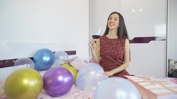 Beautiful Birthday Woman with Red Dress Sitting on Bed with Balloons and Holding a Glass of Red Wine alt