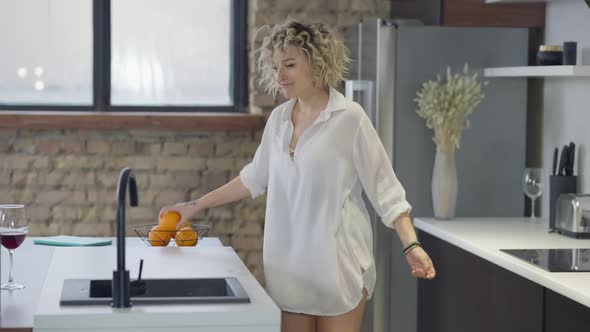 Relaxed Happy Slim Woman Dancing in Kitchen Juggling Orange and Looking at Camera Smiling alt