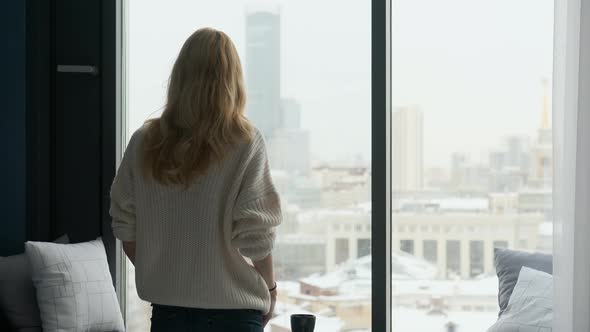 blonde woman stands with her back and looks out the window