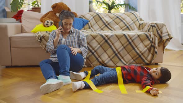 Tired Young Mother Talking on Phone Sitting on Floor Near Little Son Glued with Duct Tape alt
