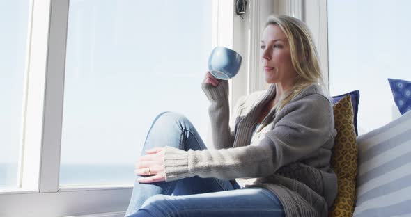 Caucasian mature woman looking through the window and drinking coffee alt