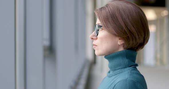 Portrait of a sad young woman with glasses standing by the window.  alt