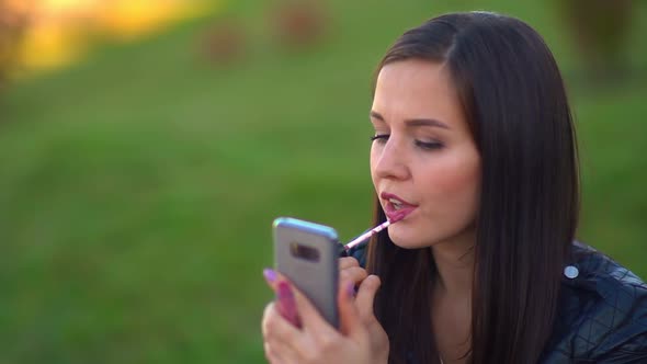 Attractive Girl in the Park Paints Her Lips, Looks at the Reflection in the Phone. Getting Ready for alt