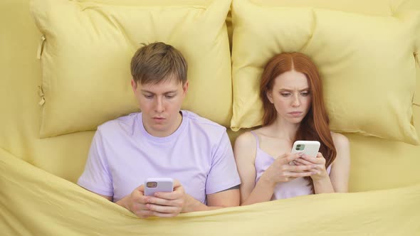 Shocked Couple Ignoring Each Other Lying in Bed at Night While Using Mobile Phones Look at Camera