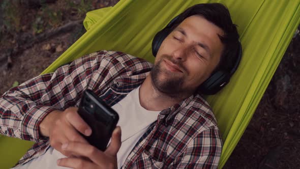 Young Caucasian Man Resting in Hammock Listening to Music on Headphones and Using Smartphone alt