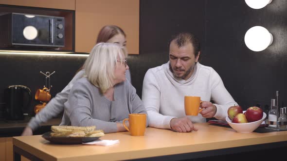 Joyful Teenage Daughter Entering Kitchen and Hugging Parents Sitting at the Table with Tea. Happy alt