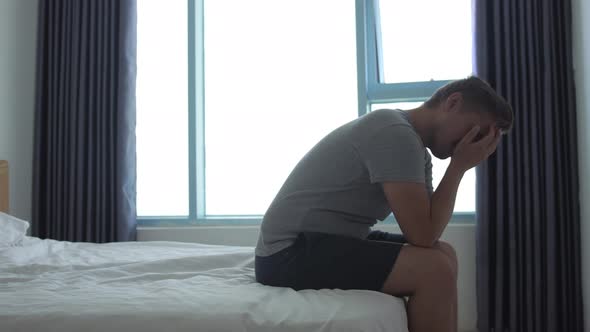 Depressed Young Man Sitting on Edge of Bed at Home or in Hotel Room