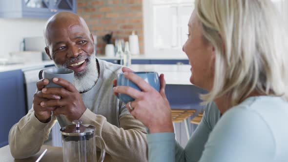 Mixed race senior couple holding coffee cups smiling looking at each other at home alt