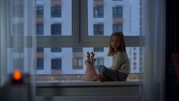 Girl Sits on Windowsill in Winter Evening and Looks Out Window at Falling Snow