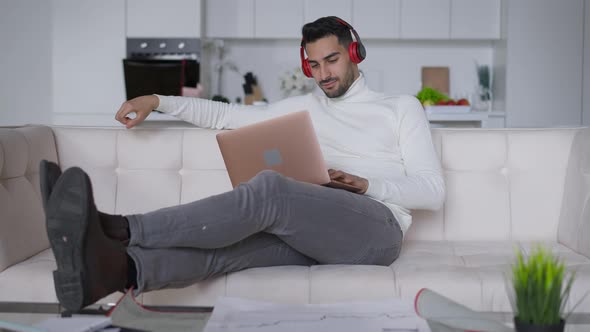 Wide Shot of Wealthy Rich Confident Young Handsome Man in Headphones Smiling Typing on Laptop alt