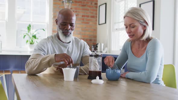 Mixed race senior couple talking to each other while having coffee at home alt