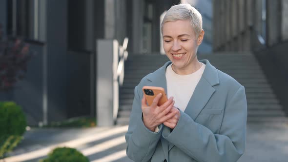 Happy Middle Aged Caucasian Woman Using Smartphone While Walking Near Business Centre alt