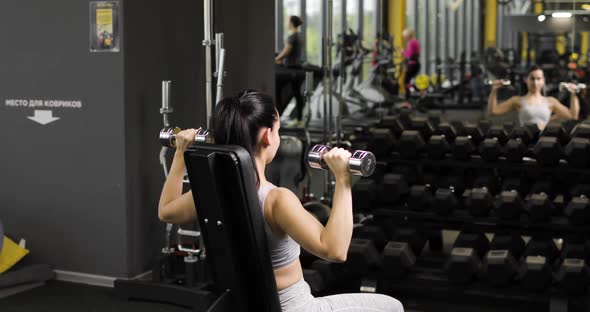 Young Brunette Woman Doing Overhead Presses with Dumbbells Sitting on a Bench in the Gym in Front of
