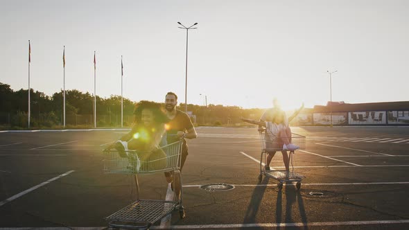 Young Multiracial Friends are Laughing and Racing on Shopping Carts at Deserted Parking Lot of alt