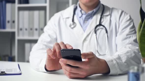 Male Doctor Uses Smartphone While Sitting At His Desk In Hospital. Symptoms And Treatment Of Disease alt