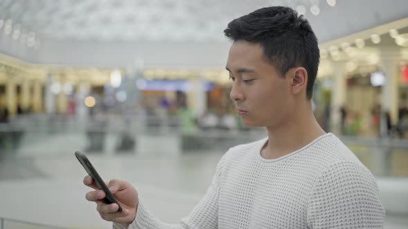 Profile Middle Shot of Asian Male Scrolling Phone on Big Mall Background