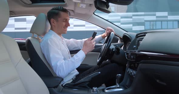 Smiling Young Man Entrepreneur Sits in the Driver's Seat in a Car and Uses a Modern Smartphone