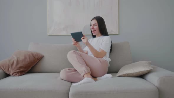 Millennial girl sitting on sofa with  tablet looking at screen in surprise.