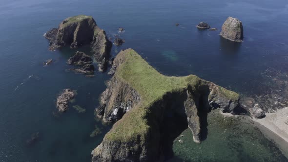 Aerial descends to reveal eroded sea caves in jagged islet in blue sea alt