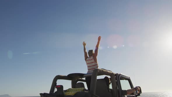 Happy caucasian gay male couple in car raising arms and waving on sunny day at the beach alt