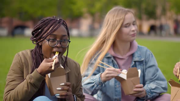 Close Up of Diverse Young Man and Women Sitting on Grass Eating Wok and Chatting alt