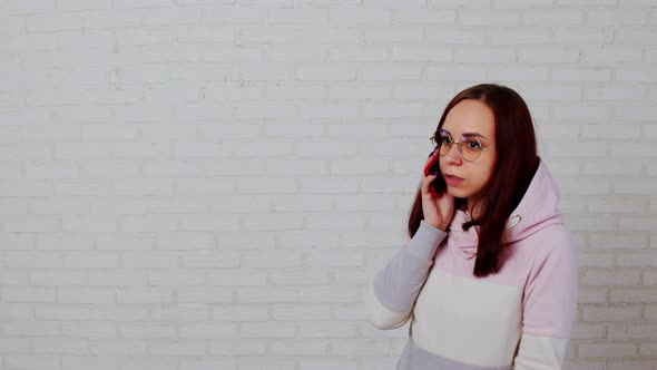 Young Woman in Glasses Speaks on Smartphone Standing Near White Brick Wall alt