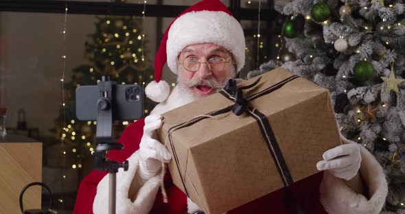 Portrait of Handsome Kind Santa Claus in His Residence Talks on Video Call Holding a Big Christmas alt