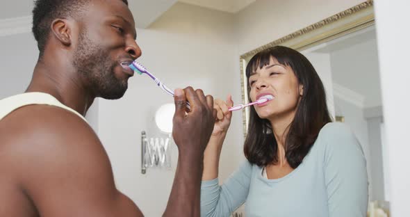Happy diverse couple looking at each other and brushing teeth in bathroom alt