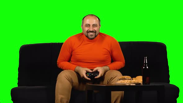Fat Man Plays the Game Console. He Is Sitting on the Couch, Drinks Beer and Eats Fast Food at Green alt