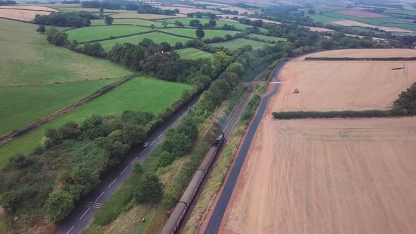 Aerial tracking forward over a Steam engineing forward out of a small station alongside a busy road alt