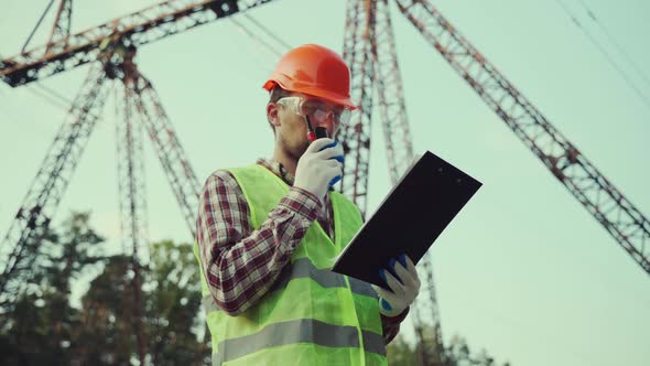Caucasian Engineer in Protective Uniform and Orange Hard Hat Holds Walkietalkie and Clipboard on alt