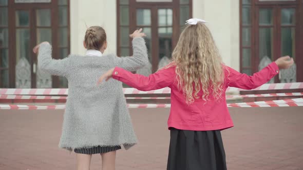 Back View of Schoolgirl Waving To School Closed During Covid-19 Pandemic. Middle Shot of Two Girls alt